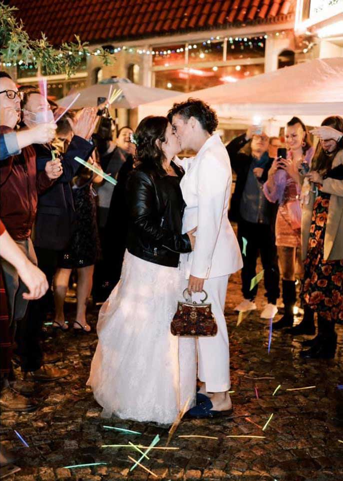 richmond-courtney-paige A couple kisses outdoors at night, surrounded by people celebrating with glow sticks and confetti. The bride wears a white dress and black jacket, and the partner wears a white suit.