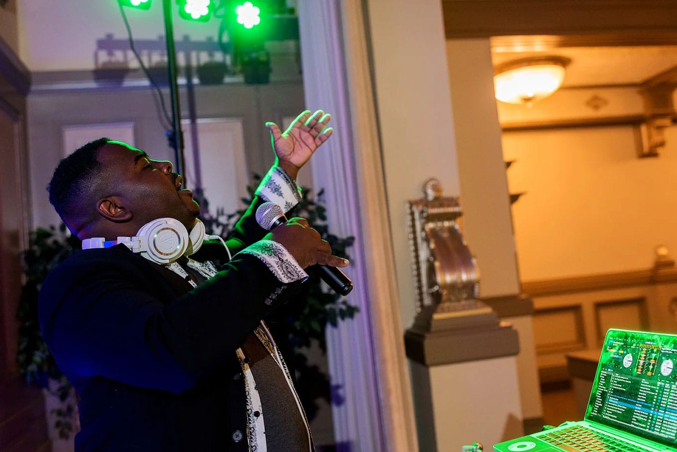 A DJ holds a microphone and gestures with one hand while standing near DJ equipment, green stage lights shining above.