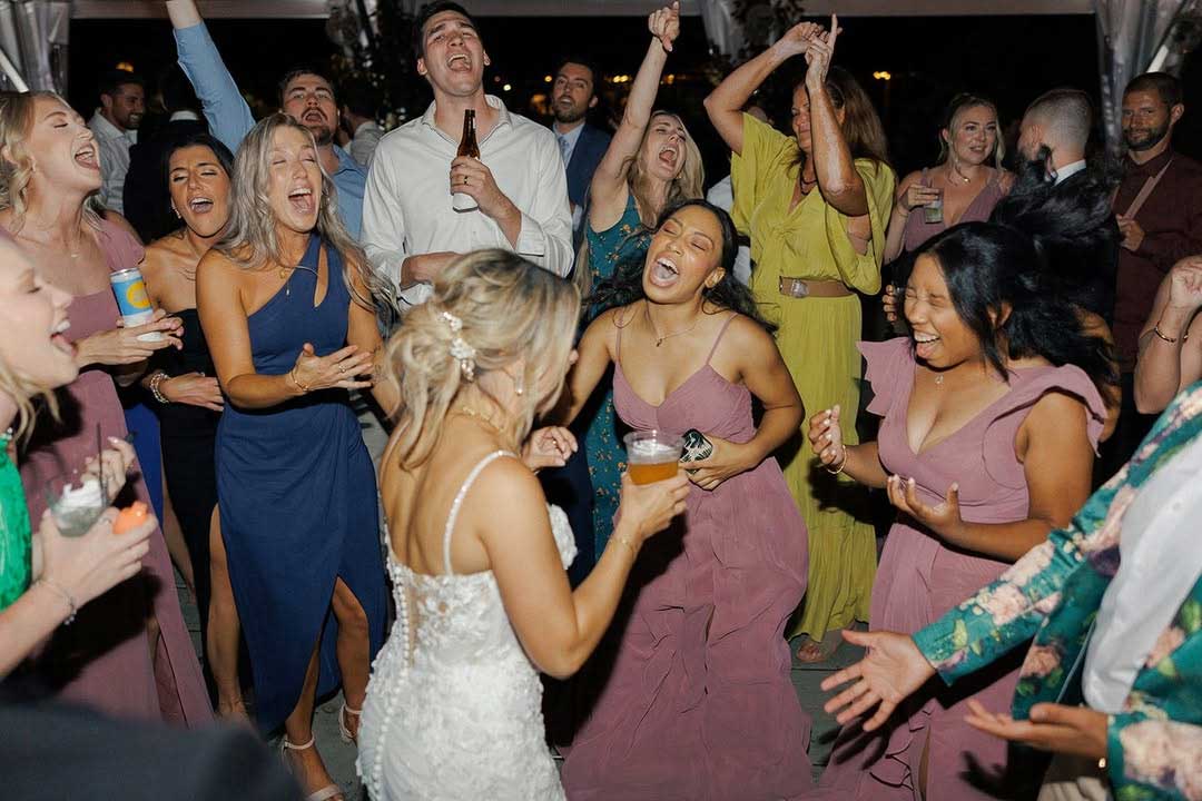 richmond-dj-girls-on-dance-floor A bride in a white dress dances with guests, who are laughing and cheering at a lively indoor event. Several people hold drinks and appear to be enjoying the celebration.