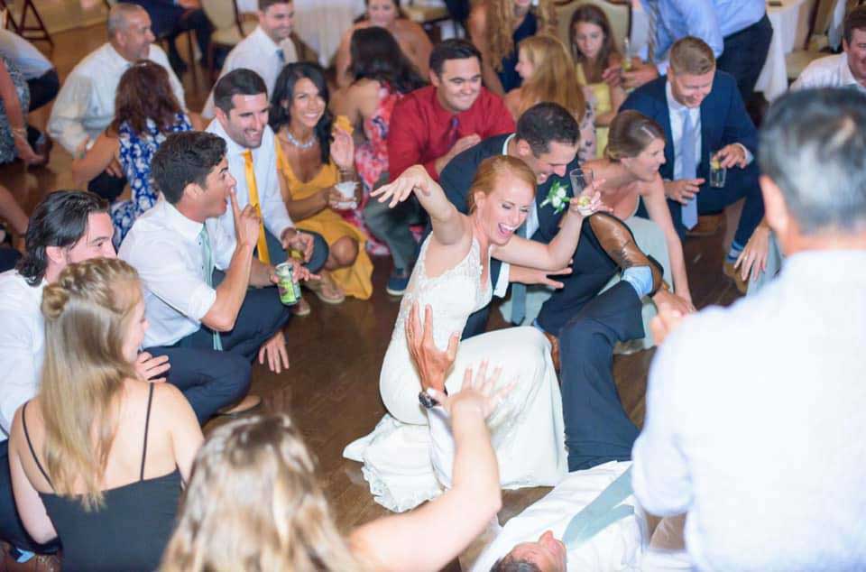 richmond-morgan-james A group of people, many in formal attire, sit and kneel on a wooden floor at a lively indoor event, with a woman in a white dress reaching toward a person lying down.