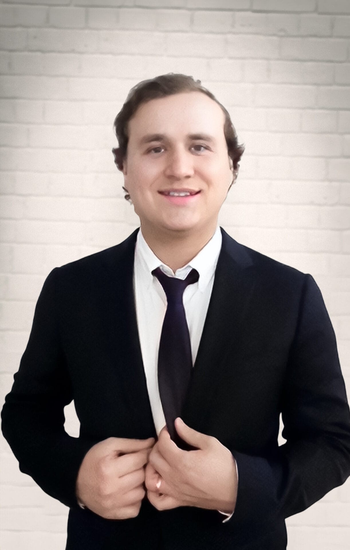 A man in a dark suit and tie stands against a white brick wall, smiling and buttoning his jacket.