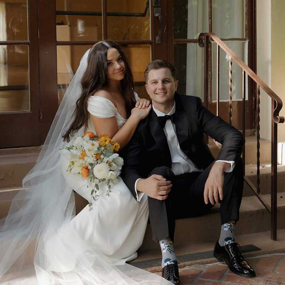 A bride in a white gown and veil sits beside a groom in a black tuxedo; she holds a bouquet of flowers and both are smiling on a staircase.