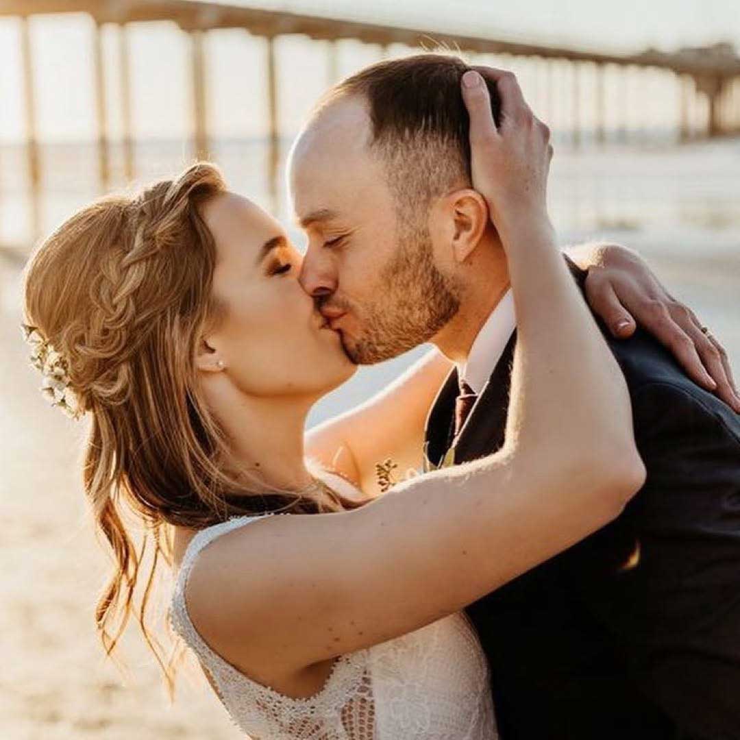 A bride and groom kiss on a beach, with a pier visible in the background and sunlight illuminating the scene.