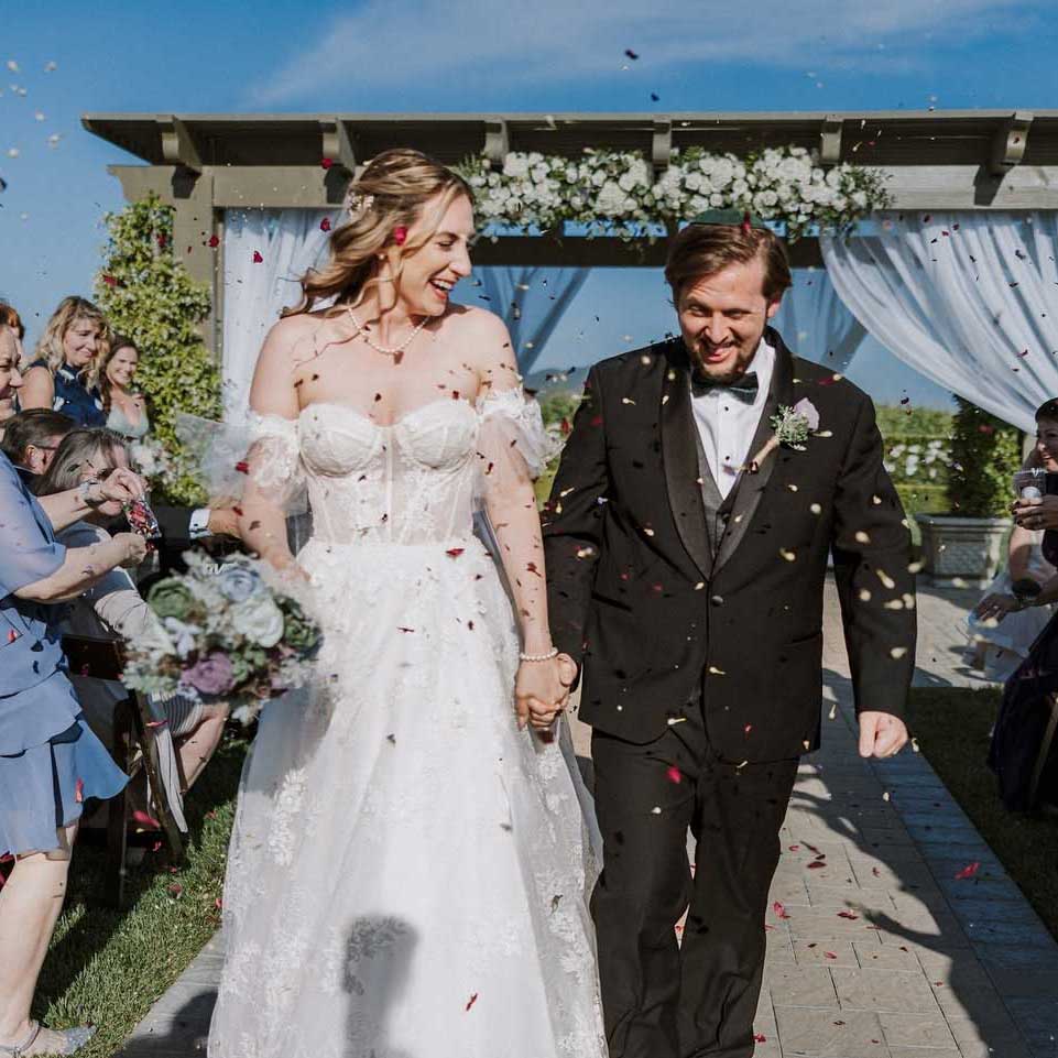 A bride and groom walk down the aisle outdoors, holding hands and smiling, as guests throw flower petals.