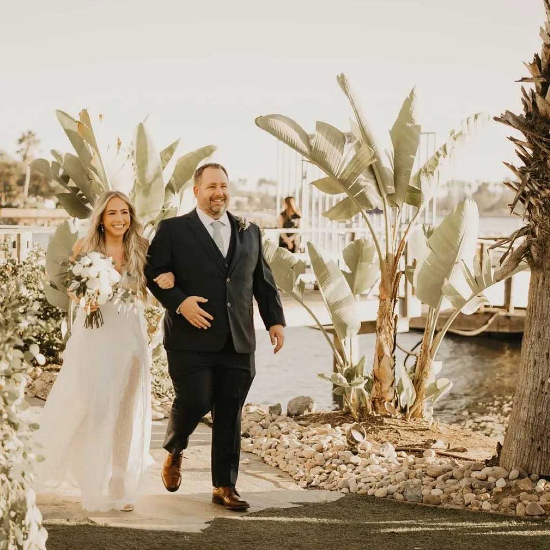 A bride in a white dress walks arm-in-arm with a man in a dark suit along a path by the water, surrounded by tropical plants.