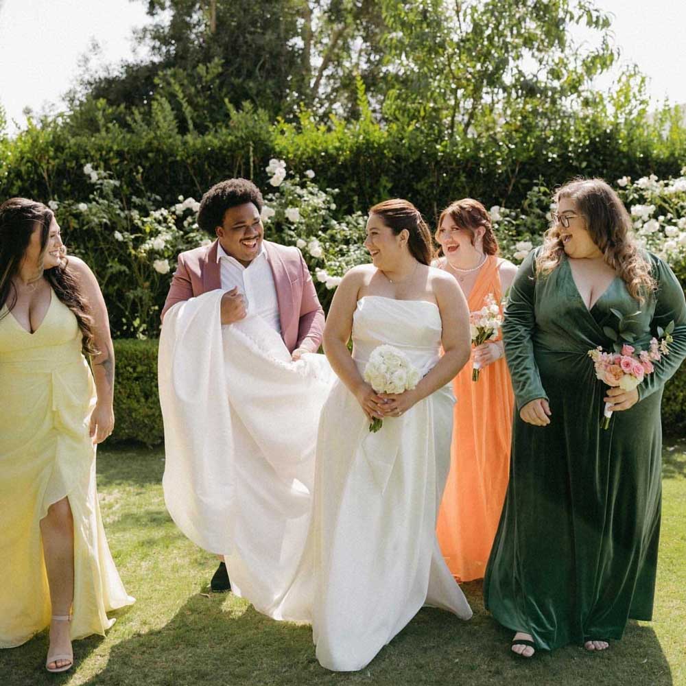 Five people dressed in formal attire, including a bride in a white dress, walk together outside on grass, smiling and holding bouquets.