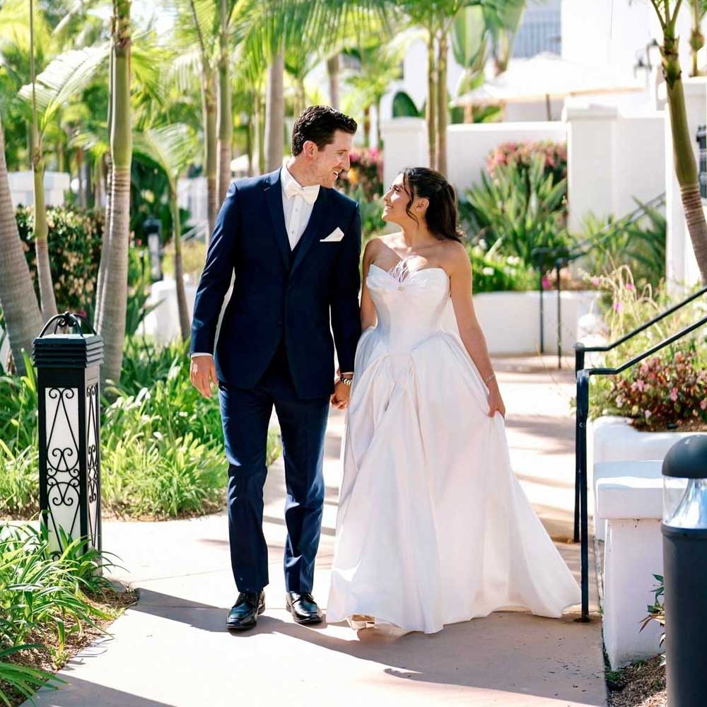 A man in a navy suit and a woman in a white wedding dress walk hand-in-hand outdoors on a sunny day, surrounded by greenery and palm trees.