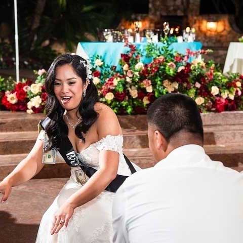 A bride wearing a white dress and sash with money pinned to it smiles and interacts with a seated guest at an outdoor event with floral decorations.