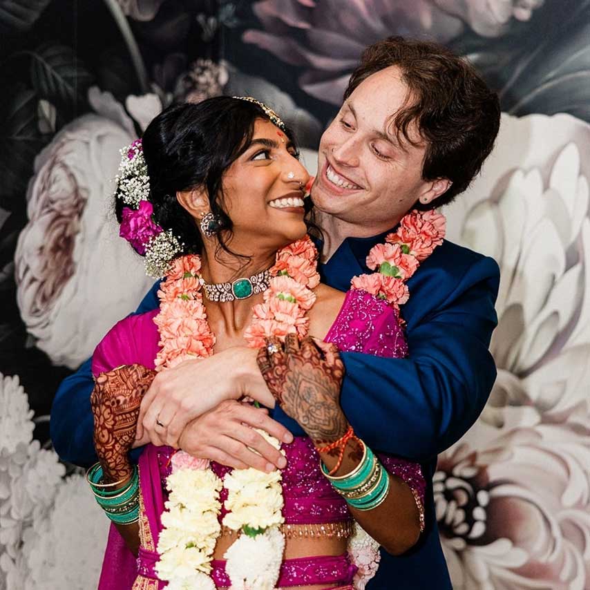 A couple in traditional attire, adorned with floral garlands, embraces and smiles at each other against a floral backdrop.