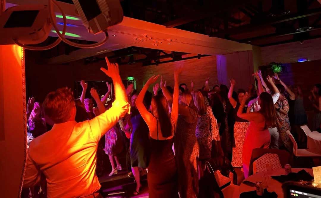 san-diego-dj-dance-floor-lighting A group of people dance with raised arms under colorful lighting at an indoor party or event.