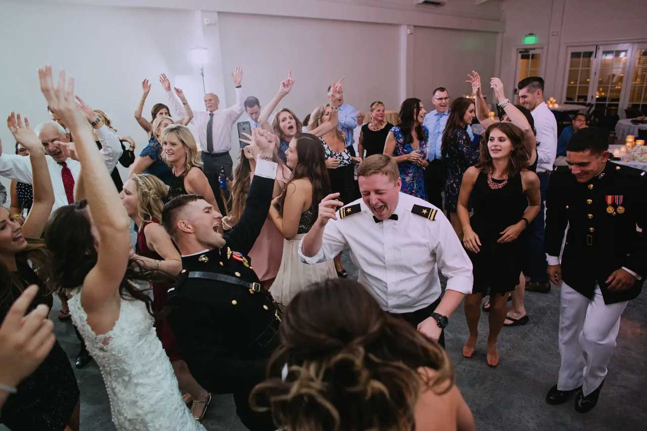 san-diego-military-wedding A group of people, including uniformed military personnel and a woman in a wedding dress, dance and celebrate at an indoor event.