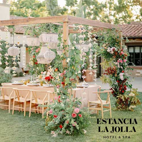 Estancia La Jolla Hotel & Spa Outdoor dining table set under a wooden pergola decorated with flowers and hanging ornaments, located at Estancia La Jolla Hotel & Spa.