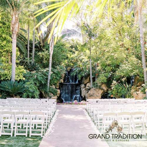 Grand Tradition Estate & Gardens Rows of white chairs face a waterfall surrounded by lush greenery at an outdoor event space. "Grand Tradition Estate & Gardens" is labeled in the bottom right corner.