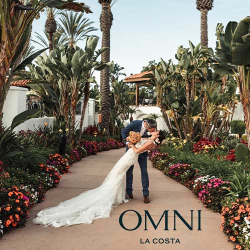 Omni La Costa A bride and groom kiss in a landscaped garden walkway surrounded by flowers and palm trees at Omni La Costa.