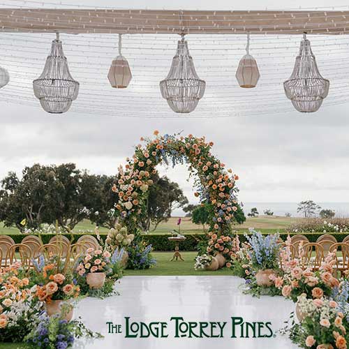 The Lodge at Torrey Pines Outdoor wedding ceremony setup with a floral arch, flower arrangements, chandeliers, and rows of chairs; ocean view in the background. Text reads: "The Lodge Torrey Pines.