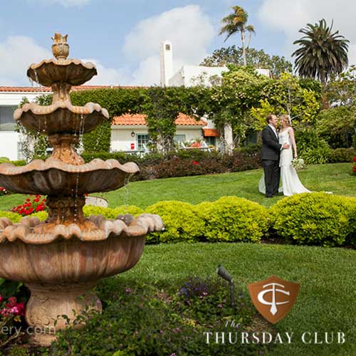 The Thursday Club A bride and groom stand on a green lawn near a large tiered fountain, with a white building and gardens in the background. "The Thursday Club" logo appears in the corner.