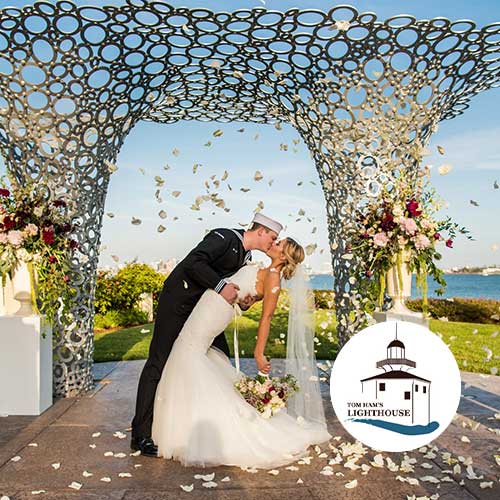 Tom Ham’s Lighthouse A bride and groom kiss under a decorative outdoor arch by the water, with flower petals falling. Tom Ham’s Lighthouse logo is in the lower right corner.