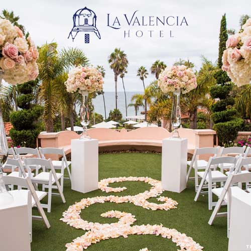 La Valencia Hotel Outdoor wedding setup at La Valencia Hotel with white chairs, floral arrangements, and a flower petal aisle, overlooking the ocean and palm trees.