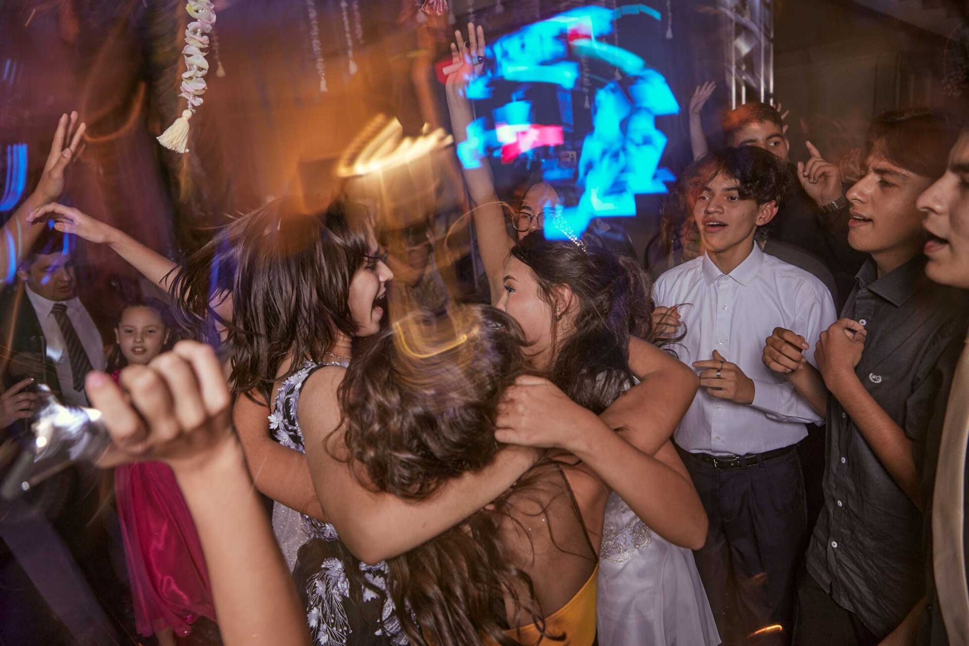 school-dance-quinceanera A group of teenagers dance and celebrate closely together at an indoor party, with colorful lighting and blurred motion conveying excitement.