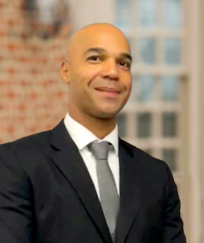 tony-nunez A man in a black suit, white shirt, and gray tie stands indoors in front of a blurred background featuring a window and brick wall.
