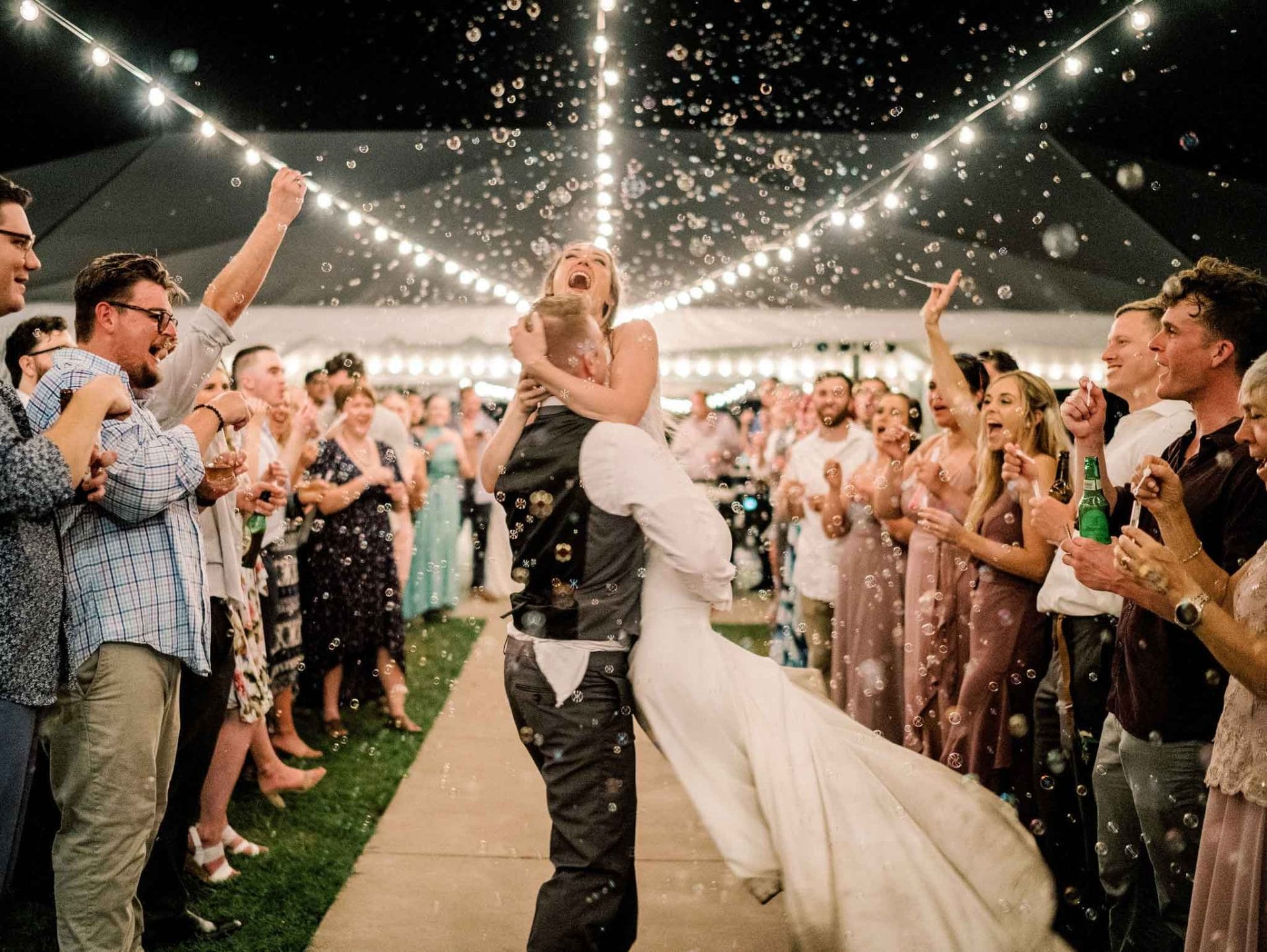 A bride and groom dance under string lights, surrounded by guests blowing bubbles and cheering at an outdoor nighttime wedding reception.
